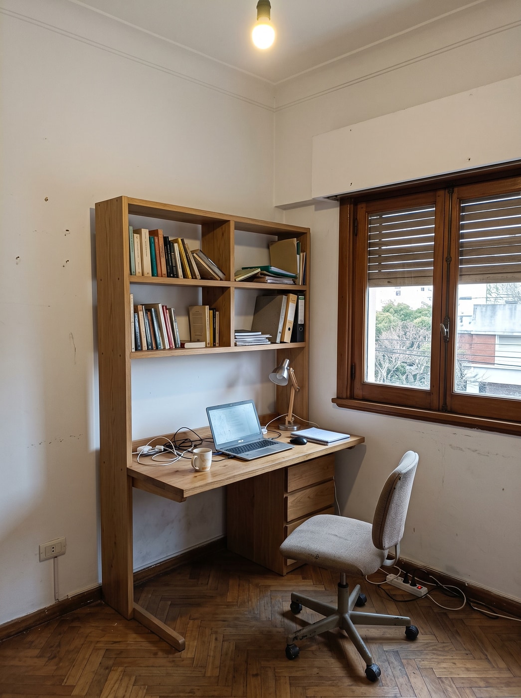 Biblioteca de roble con escritorio integrado y cajones, con laptop, lámpara y silla, junto a una ventana con postigos de madera.