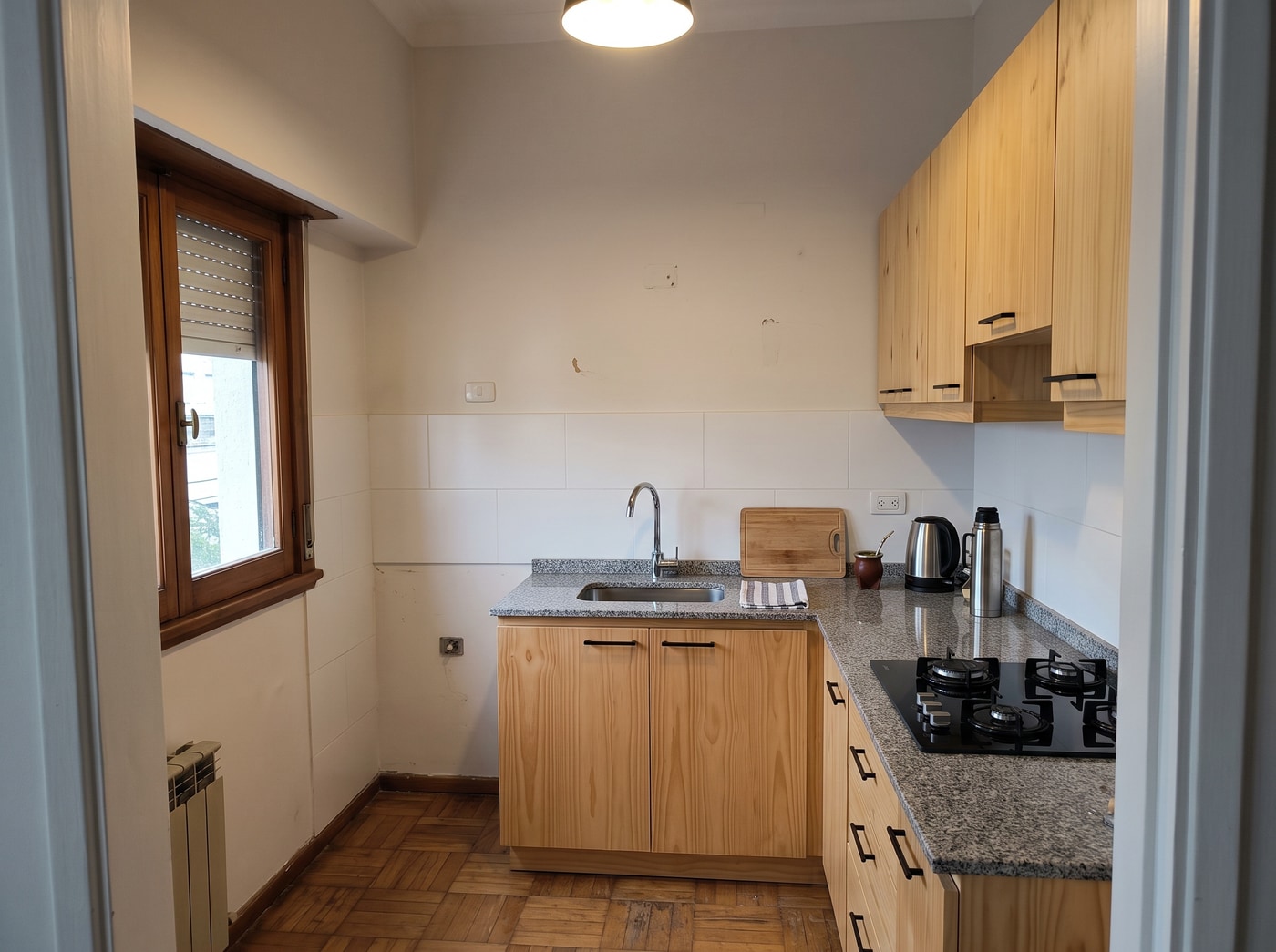 Cocina renovada con bajo mesada y alacena de pino macizo de frentes lisos, mesada de granito gris y cocina a gas, en un apartamento de Montevideo.