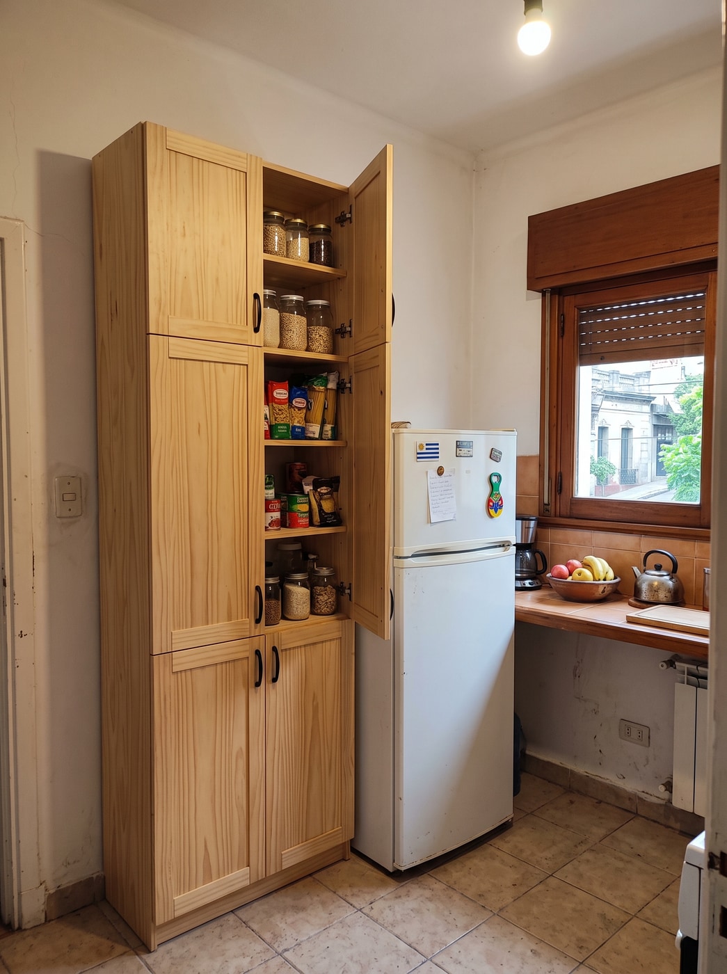 Despensero alto de pino macizo de piso a techo con una puerta abierta, en un rincón de cocina junto a la heladera.