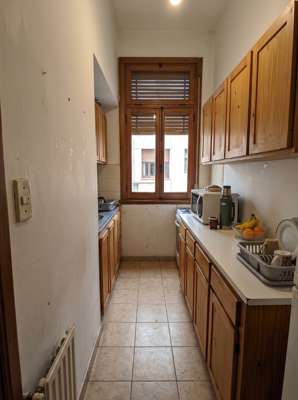 Cocina angosta tipo pasillo con muebles de pino a ambos lados, piso cerámico y ventana con postigos al fondo.