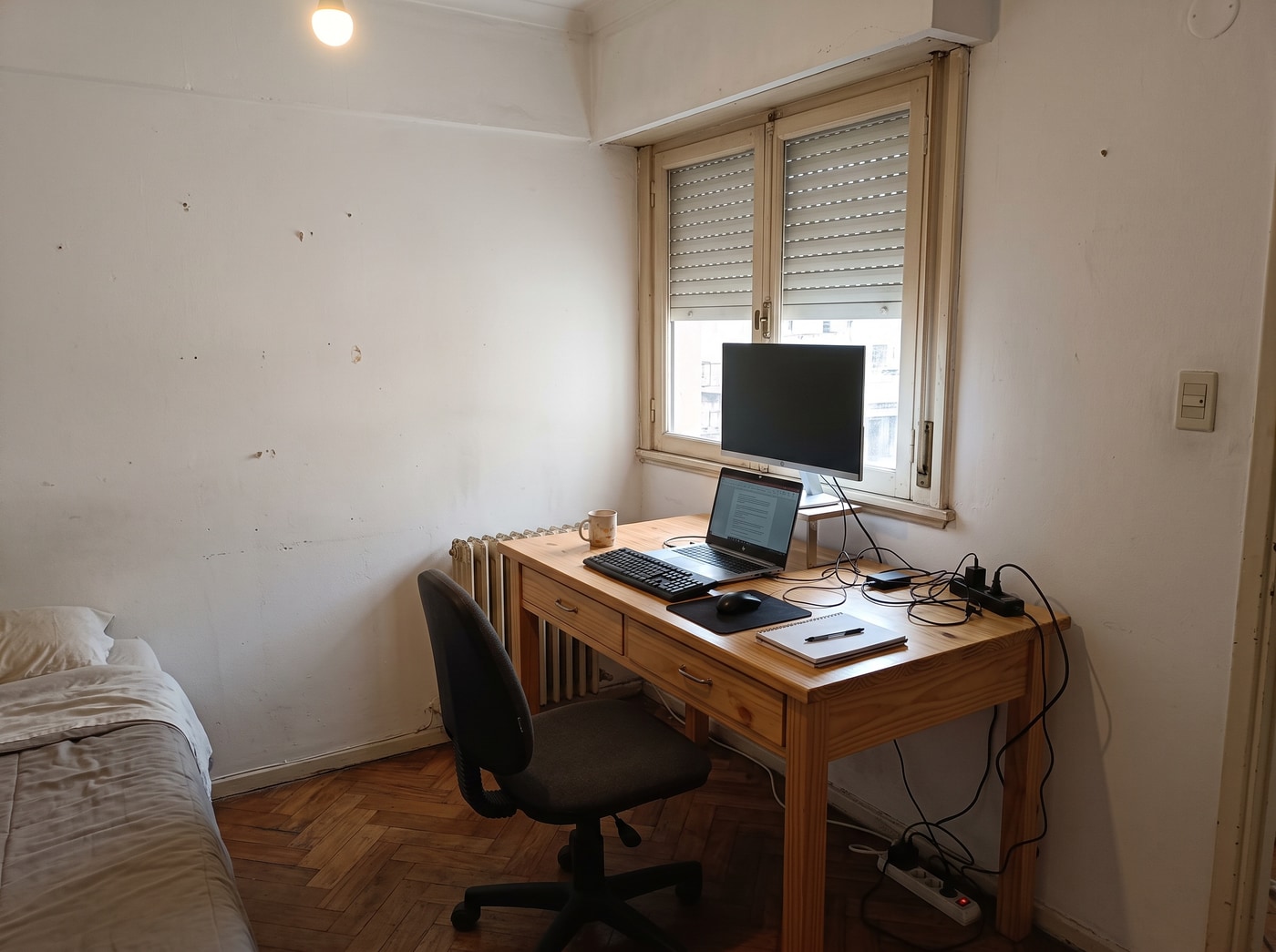 Escritorio de pino macizo con dos cajones bajo la ventana de un dormitorio, con monitor, laptop y silla de oficina.
