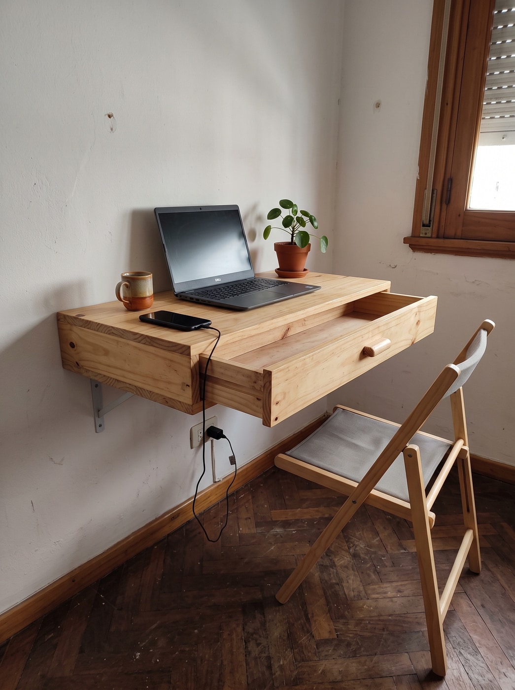 Escritorio flotante de pino macizo con un cajón, fijado a la pared de un dormitorio, con laptop y celular cargando.