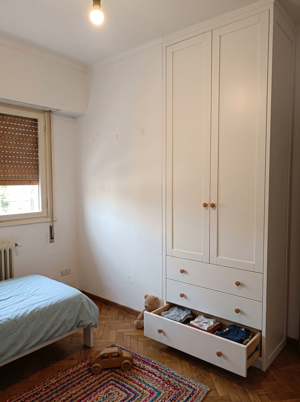 Placard infantil laqueado blanco con dos puertas y tres cajones inferiores, uno abierto, con juguetes en el piso del cuarto.